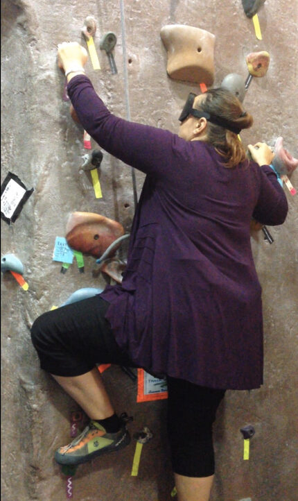 I am wearing a blindfold while climbing a indoor rock wall. My eyesight is unreliable  so I trust my foot and hand placement.  “I do things only I do them differently”. Gina Martin 