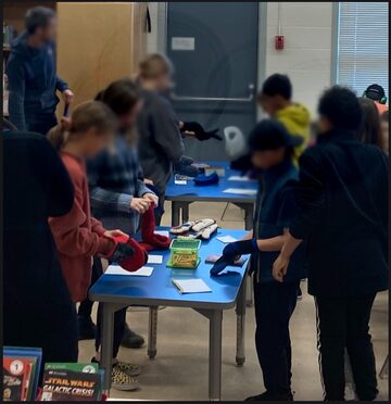 Students are standing around a table and are putting on gloves, mittens,or oven mitts to accomplish a writing task. The teacher is at the left, towards the back, watching the students participate.
