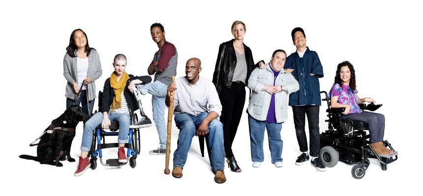 A group of people, some with apparent disabilities, 1 with a guide dog, 1 sitting down on their walker, 1 in a wheelchair, 1 with a walking stick, 1 with a prosthetic leg, and the others may or may not have apparent disabilties.