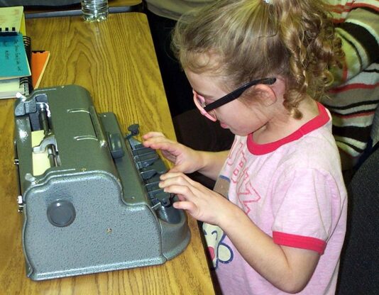 Child with ponytail, pink glasses and pink t-shirt, using Perkins Brailler Braille writer