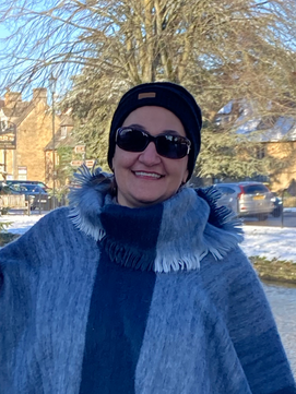 Gina is smiling at the camera. She's wearing sunglasses, a soft fuzzy purple cape and a burgundy touque, it's winter with a bit of snow on the ground behind her.