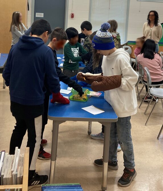 Students trying hands on activities to gain a better understanding about disabilities. The teacher is at the back, on the right, watching the students participate.